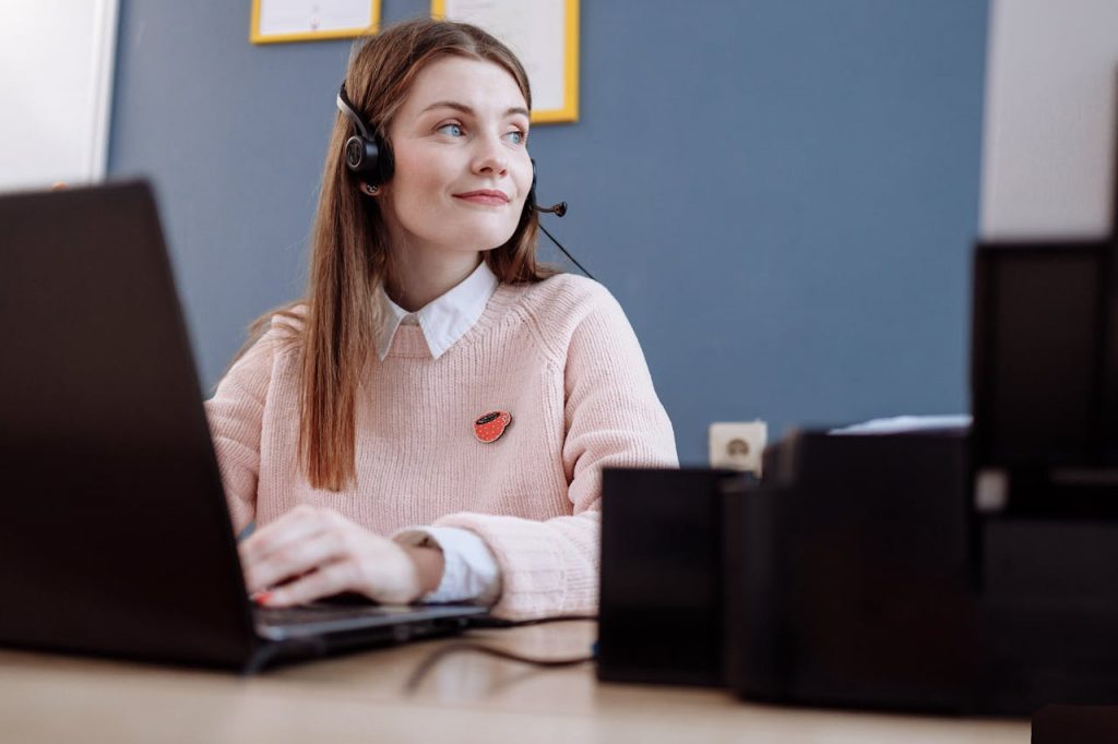 pexels-photo-6502999 A cheerful woman in a pink sweater working remotely with a laptop and headset, providing customer service.